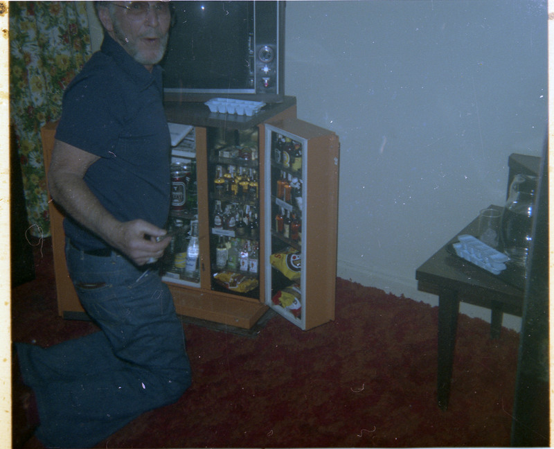 A photograph of Donald Crabtree posing in front of a hotel mini fridge. The mini fridge is filled with various beverages and snacks. A television and coffee maker are also visible.