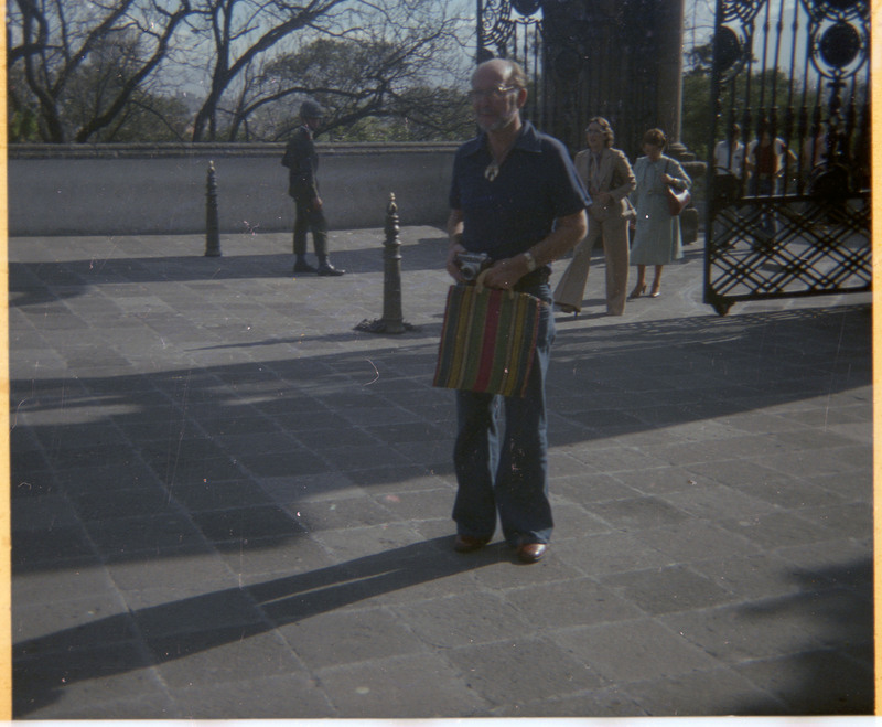 A photograph of Donald Crabtree standing in front of a gate and looking in the distance. Crabtree is holding a camera and a bag.