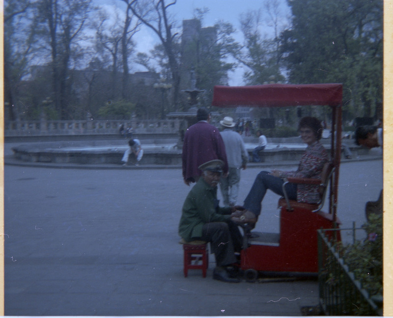 A photograph of Evelyn Crabtree getting her shoes shined. A found and other buildings are visible in the distance.