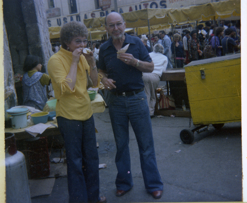 A photograph of Donald and Evelyn Crabtree eating food prepared by street vendors. An unnamed woman preparing food and a crowd of people are visble behind them.