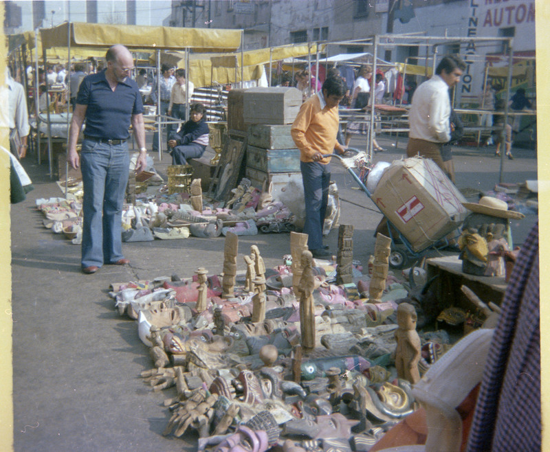 A photograph of Donald Crabtree looking at a street market stall. The stall in the photograph appears to be selling sculptures and masks made of unnamend materials.