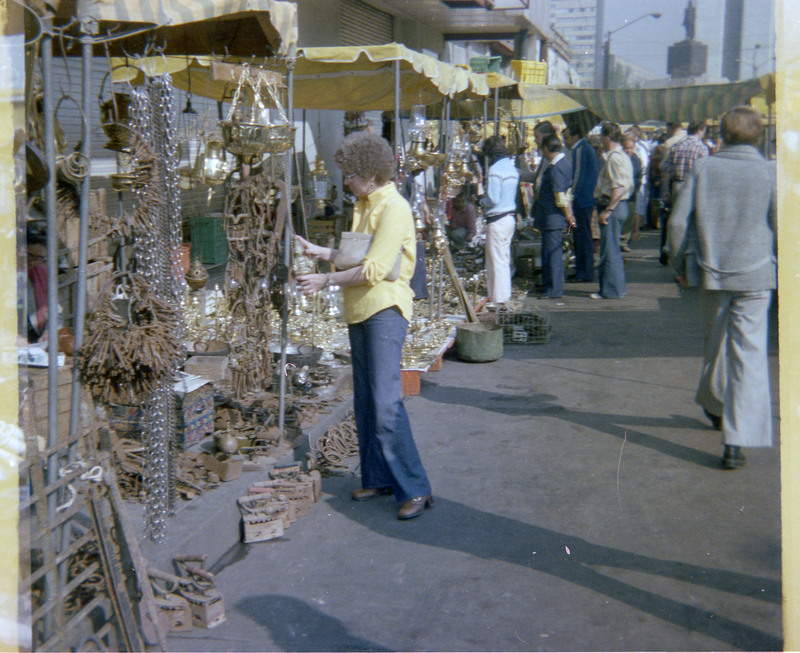 A photograph of Evelyn Crabtree looking at a street market stall. The stall in the photograph appears to be selling various types of metal items.