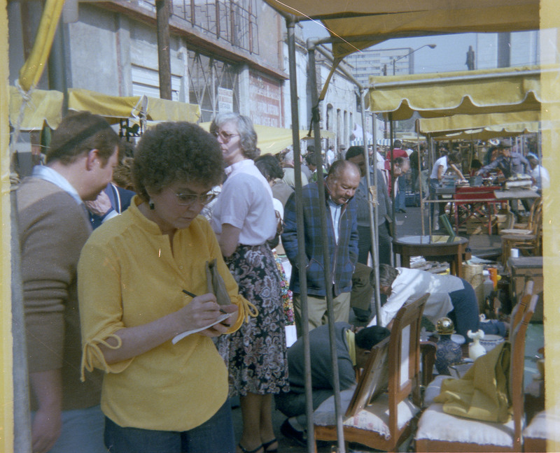 A photograph of Evelyn Crabtree looking at a street market stall. The stall in the photograph appears to be selling furniture items and Crabtree is writing notes.