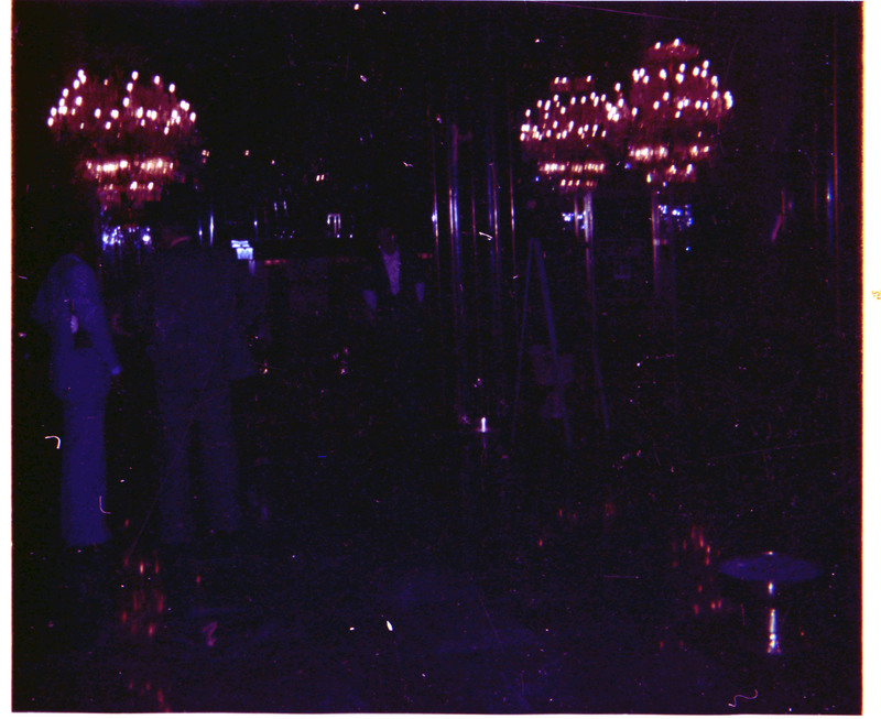 A photo of a dark event space with chandelier lights. There appear to be at least three people in the image.