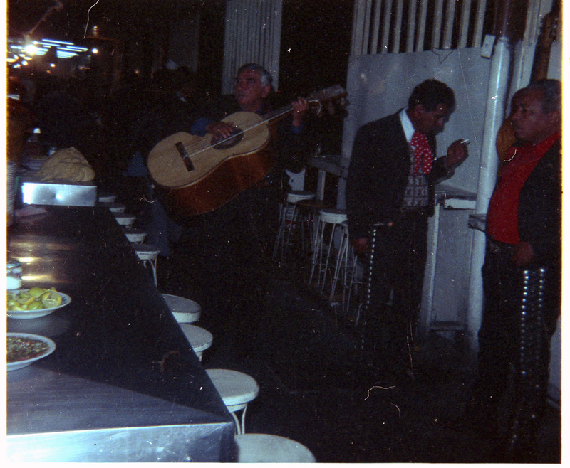 A photo of members of a band, with one playing a guitar. A table of food is visible on the left side of the image.