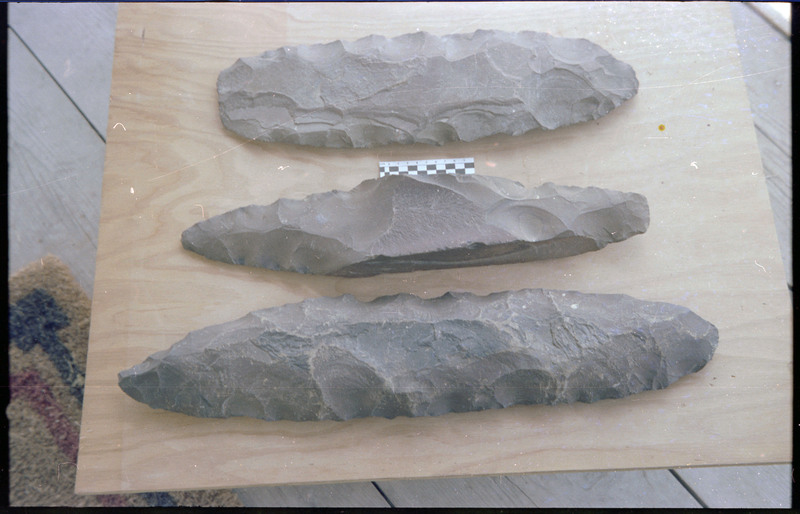 A photo of three flintknapped pieces on a piece of wood. A small scale bar is visible between the top two pieces. Associated item numbers: ce_b83_f2-item4-025 through ce_b83_f2-item4-028.