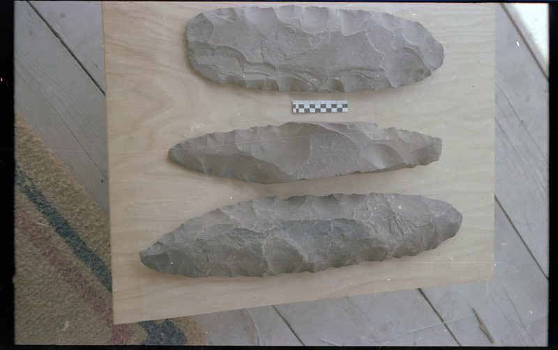 A photo of three flintknapped pieces on a piece of wood. A small scale bar is visible between the top two pieces. Associated item numbers: ce_b83_f2-item4-025 through ce_b83_f2-item4-028.