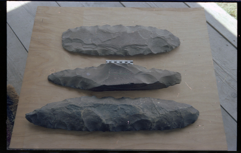 A photo of three flintknapped pieces on a piece of wood. A small scale bar is visible between the top two pieces. Associated item numbers: ce_b83_f2-item4-025 through ce_b83_f2-item4-028.