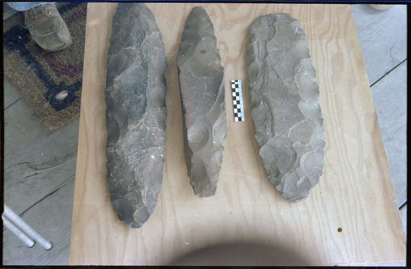 A photo of three flintknapped pieces on a piece of wood. A small scale bar is visible between the top two pieces. Associated item numbers: ce_b83_f2-item4-025 through ce_b83_f2-item4-028.