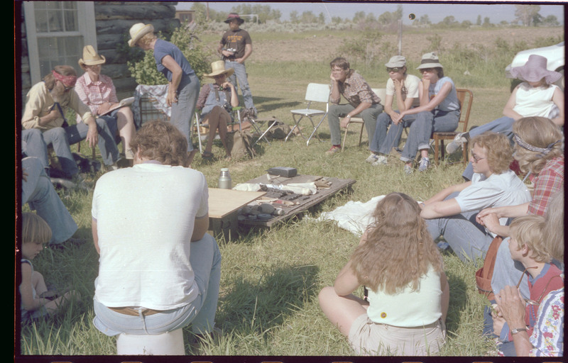 A group of people sitting outside around a ground-level table. The table appears to have different tools and materials. Associated item numbers: ce_b83_f2-item4-029 and ce_b83_f2-item4-031 through ce_b83_f2-item4-042.