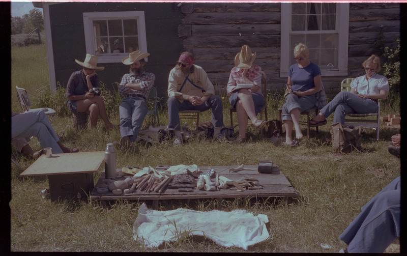 A group of people sitting outside around a ground-level table. The table appears to have different tools and materials. Associated item numbers: ce_b83_f2-item4-029 and ce_b83_f2-item4-031 through ce_b83_f2-item4-042.