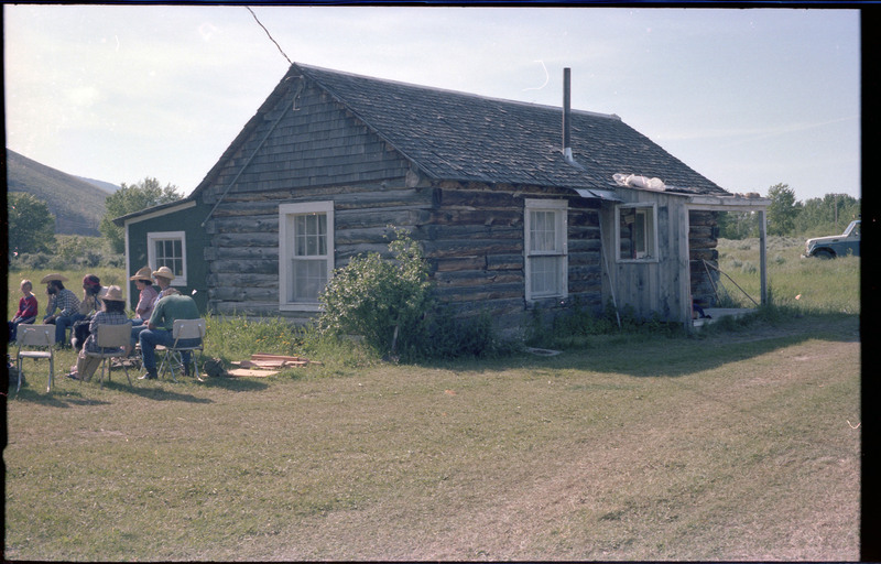 A photo of a group of people sitting outside with a house in the foreground. A vehicle is also visible in the distance. Associated item numbers: ce_b83_f2-item4-029 and ce_b83_f2-item4-031 through ce_b83_f2-item4-042.