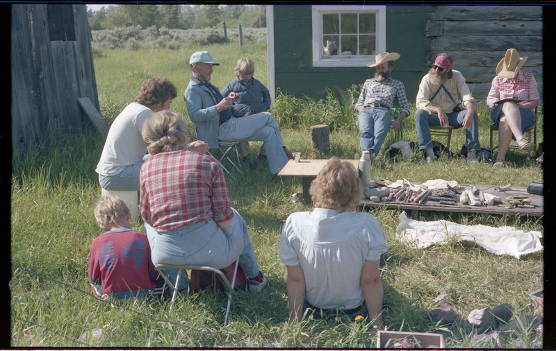 A photo of Donald Crabtree talking with a group of people sitting outside around a ground-level table. The table appears to have different tools and materials. Associated item numbers: ce_b83_f2-item4-029 and ce_b83_f2-item4-031 through ce_b83_f2-item4-042.