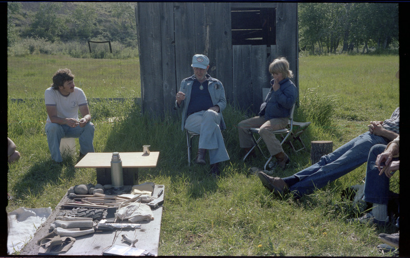 A photo of Donald Crabtree talking with a group of people sitting outside around a ground-level table. The table appears to have different tools and materials. Associated item numbers: ce_b83_f2-item4-029 and ce_b83_f2-item4-031 through ce_b83_f2-item4-042.