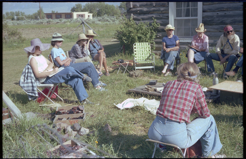 A group of people sitting outside around a ground-level table. The table appears to have different tools and materials. Associated item numbers: ce_b83_f2-item4-029 and ce_b83_f2-item4-031 through ce_b83_f2-item4-042.