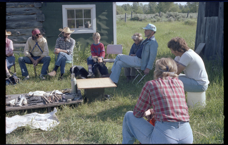 A photo of Donald Crabtree talking with a group of people, and two dogs, sitting outside around a ground-level table. The table appears to have different tools and materials. Associated item numbers: ce_b83_f2-item4-029 and ce_b83_f2-item4-031 through ce_b83_f2-item4-042.