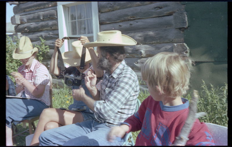 A photo of four people, including one child, sitting outside, talking with one another. One person is placing a camera around their neck. Associated item numbers: ce_b83_f2-item4-029 and ce_b83_f2-item4-031 through ce_b83_f2-item4-042.