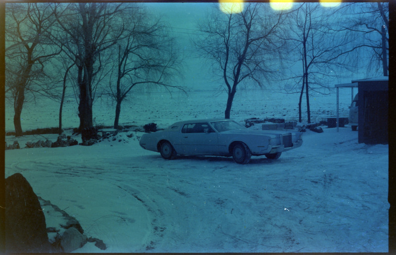 A photograph of a car parked in a snow-covered driveway. Another vehicle is visible in a carport on the right side of the screen in the background.