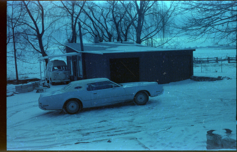 A photograph of a car parked in a snow-covered driveway. Another vehicle in a carport connected to a building is visible in the background.