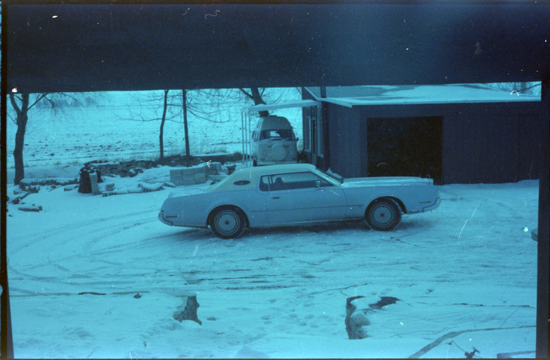 A photograph of a car parked in a snow-covered driveway. Another vehicle in a carport connected to a building is visible in the background.