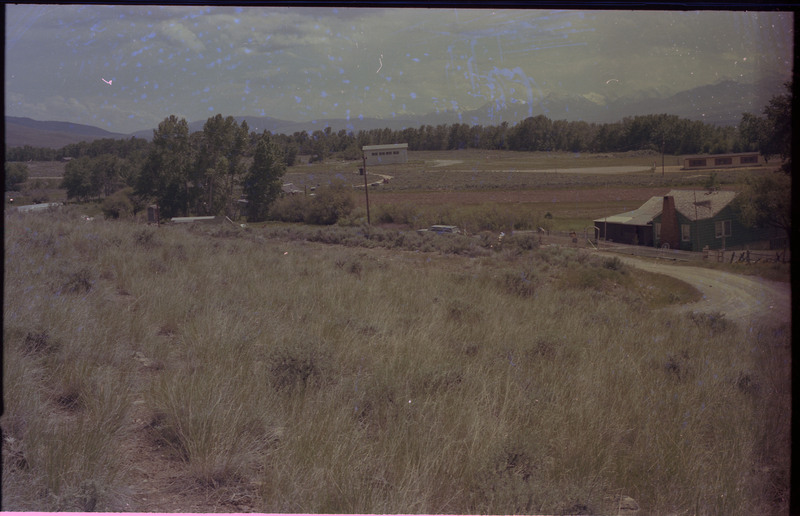 A photograph of a grassy landscape with buildings and mountains in the distance. At least one building appears to be a home.