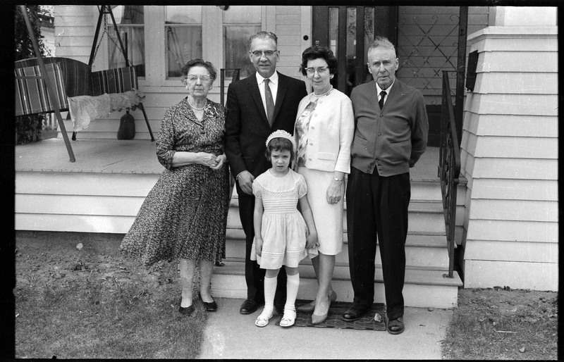 A photograph of a group of people, including one child, posing for the camera. They are standing in front of a home.