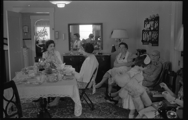 A photograph of a group of people, including one child, in a sitting or dining room. Two women are sitting at a table and appear to be conversing with each other.