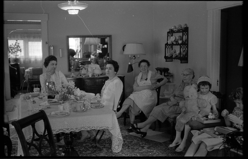 A photograph of a group of people, including one child, in a sitting or dining room. Two women are sitting at a table.