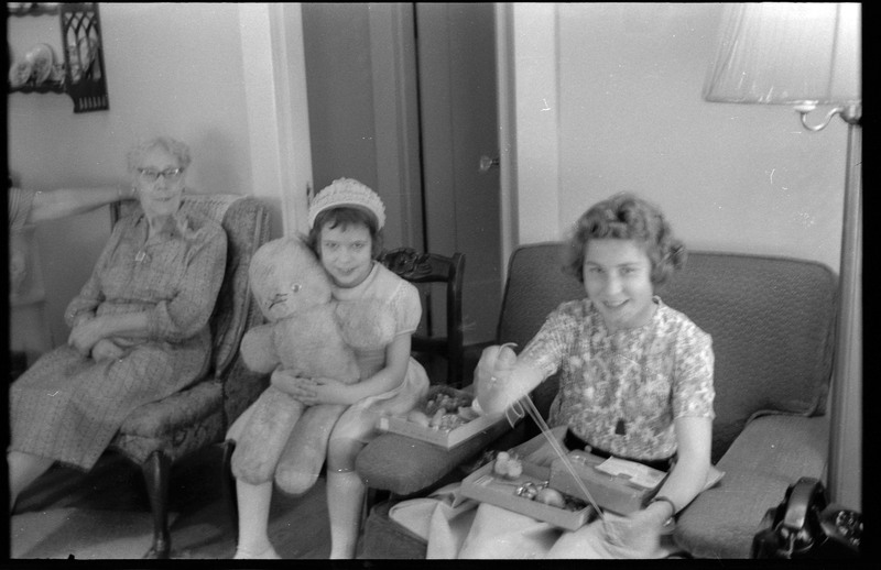 A photograph of a woman, a child, and a young woman in a sitting or dining room. The child holds a large teddy bear and the young woman holds a box.