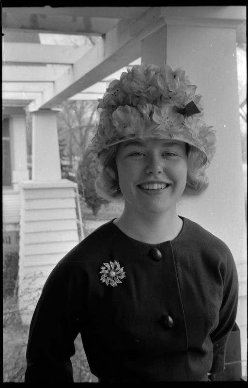 A photograph of a yound woman standing on the porch. The woman is wearing a hat that is covered in flowers.