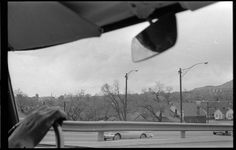 A photograph from the viewpoint of the front seat of a car. Two other vehincles are on the road and houses and other buildings are visible in the distance.