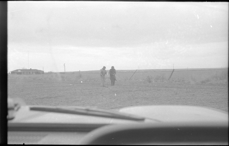 A photograph of two men standing in a field, with a house visible in the distance. It appears that the photograph was taken from inside a vehicle.