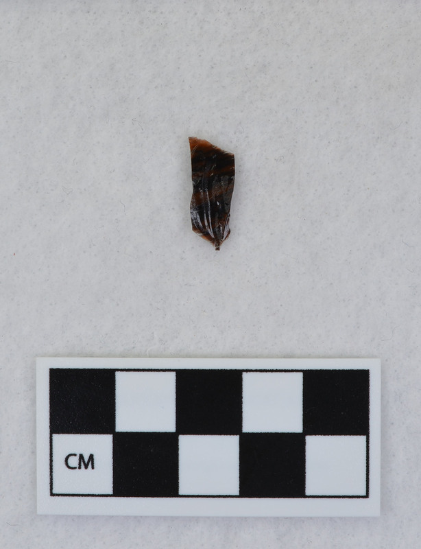 A channel flake, medial section, from a Clovis point created by Donald E. Crabtree. This object mends with seven other objects in the collection; Ce.3845 - Ce.3852. The object was made using percussion fluting with a hammerstone while the distal end rested on an anvil. Ce.3847, Ce.3848, and Ce.3849 make up the channel flake from the labeled side of the object.