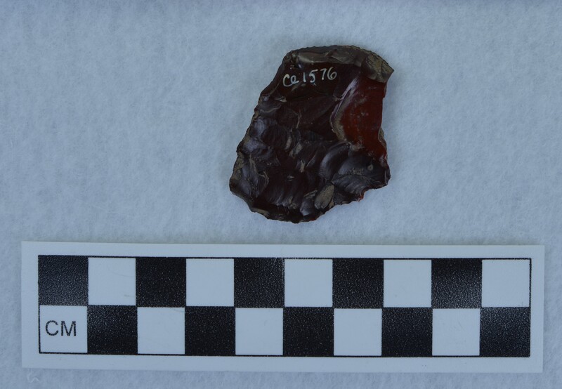 A deep red jasper projectile point, broken. It has evidence of being in fire and has pot-lidded from heat, Photo b shows: "FVII 30A" written on the artifact