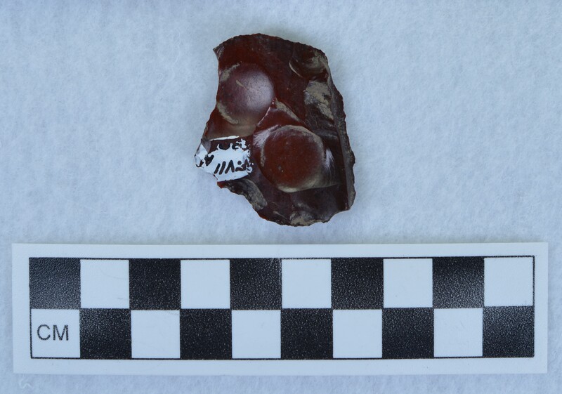 A deep red jasper projectile point, broken. It has evidence of being in fire and has pot-lidded from heat, Photo b shows: "FVII 30A" written on the artifact