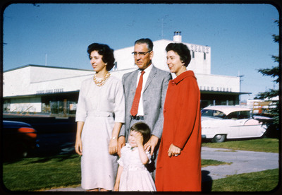 A photographic slide displaying three adults and one child standing outside of a SEARS store. From left to right there is a woman in a white dress, a man with glasses in a suit, a young girl in a white dress, and a woman in a red coat. They are facing slightly left, away from the camera.