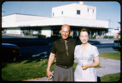 A photographic slide displaying a man and a woman standing outside a SEARS store. The man on the left is wearing a black shirt and has his arm around the woman to the right, who is wearing a white dress. They both face the camera and smile.