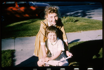 A photographic slide displaying two children smiling at the camera. A girl in an orange dress leans over a younger girl in a white dress.