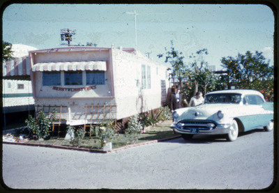 A photograph displaying a manufactured home next to a car. Slightly behind the car, there are two people. The manufactured home has a "Merry Christmas" garland.