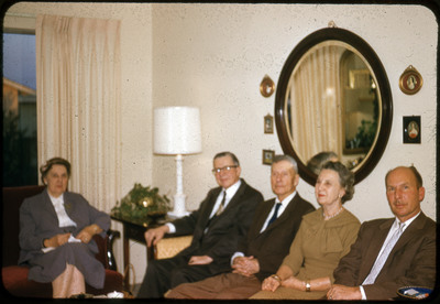 A photographic slide displaying five people sitting together inside. Two men in suits and two women in coats sit on a couch, while one woman in a jacket sits at an armchair to their left. The interior is decorated with lamps, photographs, and a mirror.