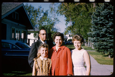A photographic slide displaying two children, a man, and a woman standing together. From left to right, there is a girl in a white dress, a woman in a red coat, a younger girl in an orange dress, and a man with glasses in a suit facing the camera. They are in front of the driveway of a house with a parked car.