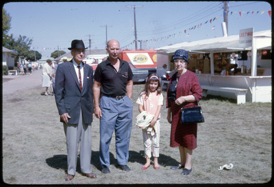 A photographic slide of two men, a woman, and a child stand next to a series of foodstands.