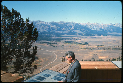 A photographic slide of a man looking at an information placard over a scenic mountainsid view.