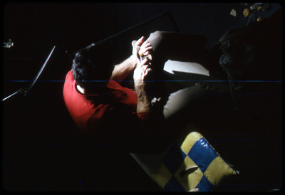 A photographic slide of a man seated while flintknapping under a bright light.