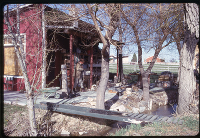 A photographic slide of a man smoking next to a red building, behind a small creek spanned by a narrow wooden bridge.