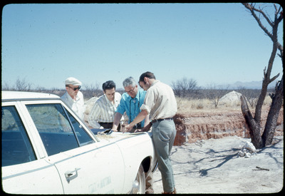 A photographic slide of four men standing over a map on the closed trunk of a car.