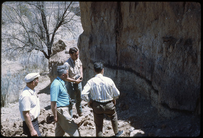 A photographic slide of four men observing the side of a rocky cliff.