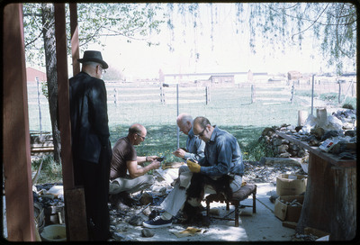A photographic slide of three men flintknapping on a porch while a fourth man watches.