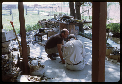 A photographic slide of two men flintknapping on a messy porch.