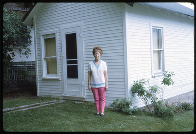 A photographic slide of a woman standing in the front yard of a white house.